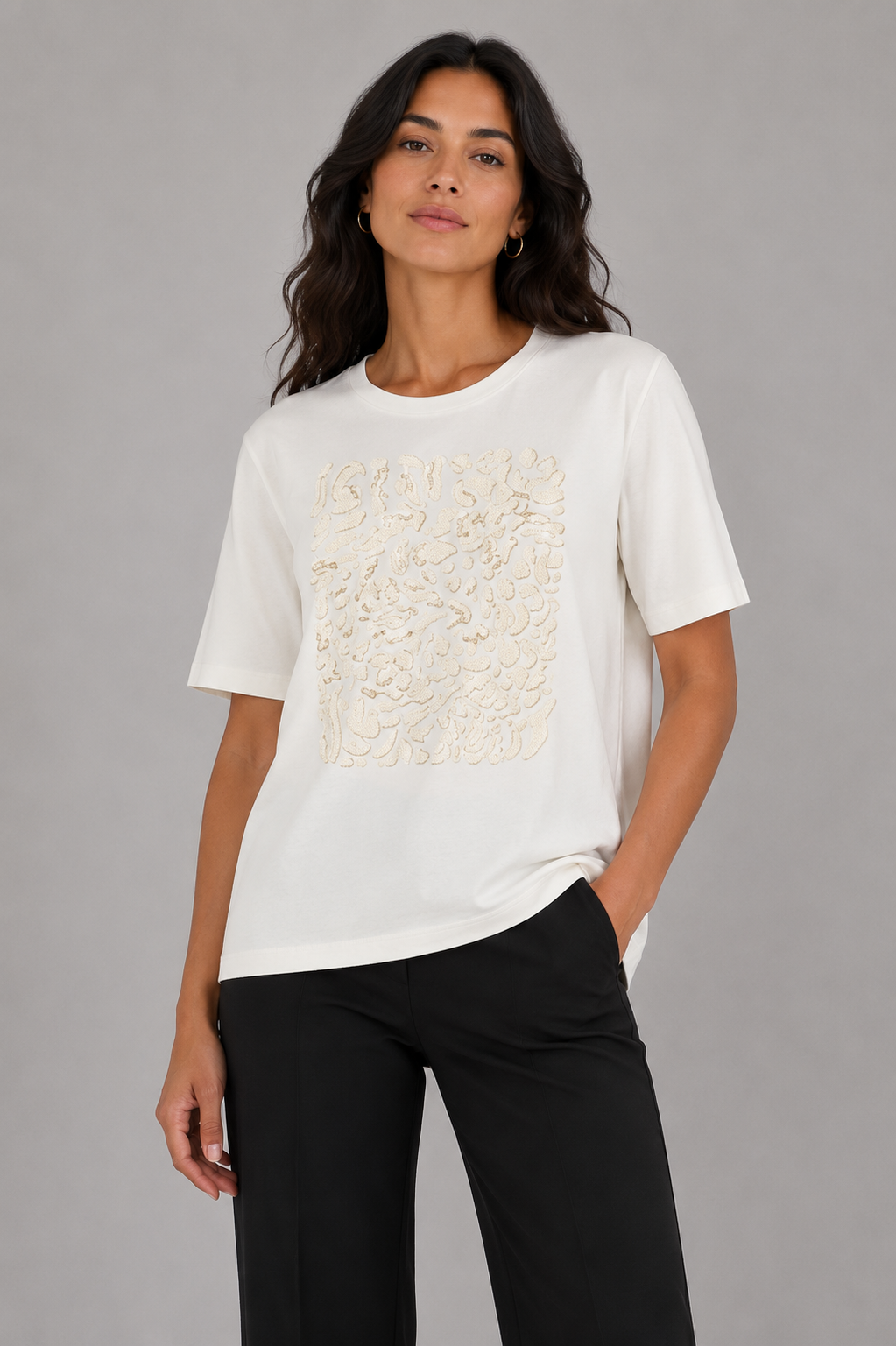 A woman modeling a white short-sleeve crewneck t-shirt with a textured beige floral lace panel on the front, paired with black pants, against a solid gray studio background.
