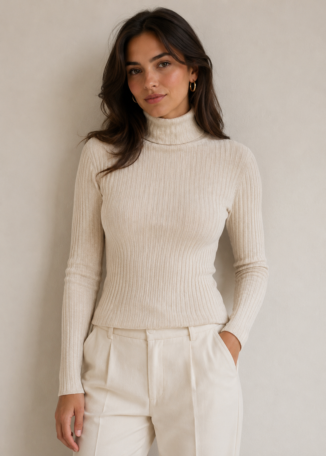 A woman wearing a fitted ribbed turtleneck sweater in warm off-white/cream, paired with matching cream trousers, standing against a plain light beige wall.
