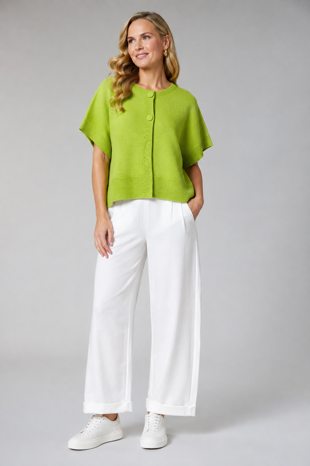 A woman modeling a bright lime green short-sleeve knit top with buttons, paired with white wide-leg trousers and white sneakers, against a neutral gray studio background.