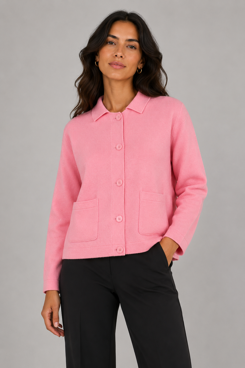 A woman modeling a light pink button-up knit jacket with a pointed collar and two front patch pockets, paired with black trousers, against a solid gray studio background.