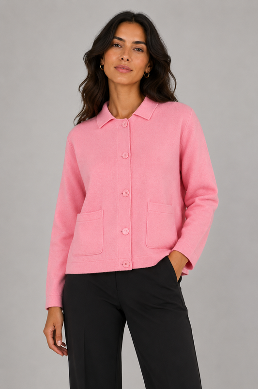 A woman modeling a light pink button-up knit jacket with a pointed collar and two front patch pockets, paired with black trousers, against a solid gray studio background.