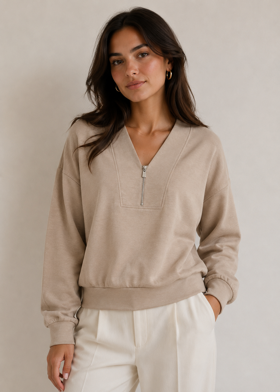A woman wearing a beige long-sleeve half-zip pullover sweater and white trousers, standing against a plain light beige studio background.