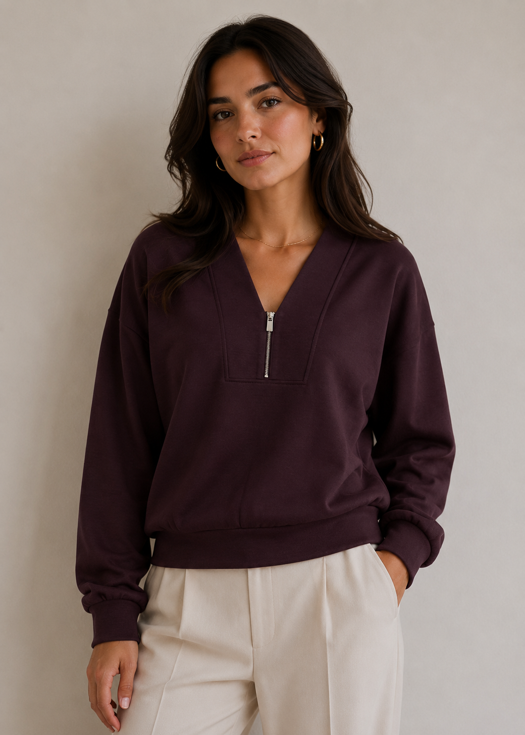 A woman wearing a dark plum purple half-zip sweatshirt with a split V-neck and silver zipper, paired with cream-colored pants, standing against a neutral light gray wall.