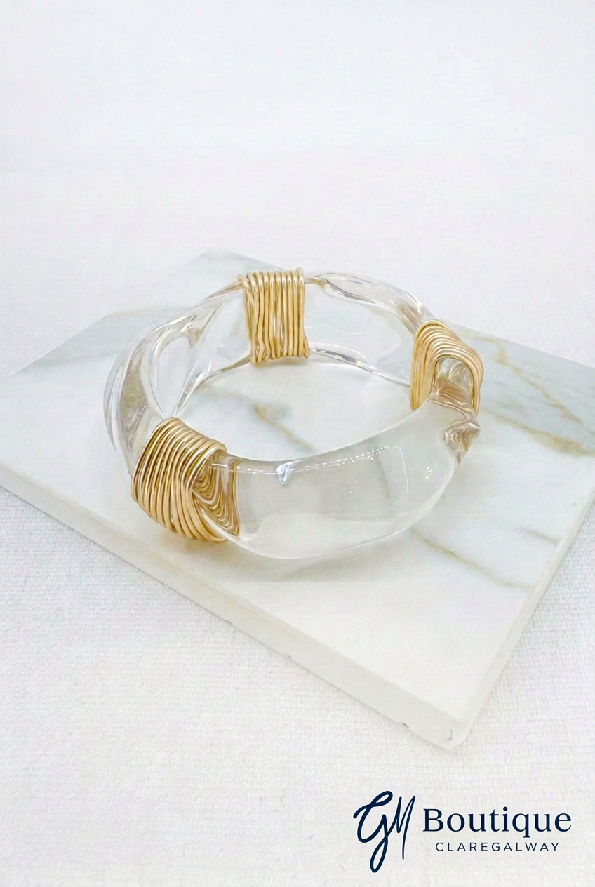 A clear acrylic or glass cuff bracelet with gold wire-wrapped ends, displayed on a white marble block against a white background. A 'GJ Boutique' watermark is visible in the bottom right corner.