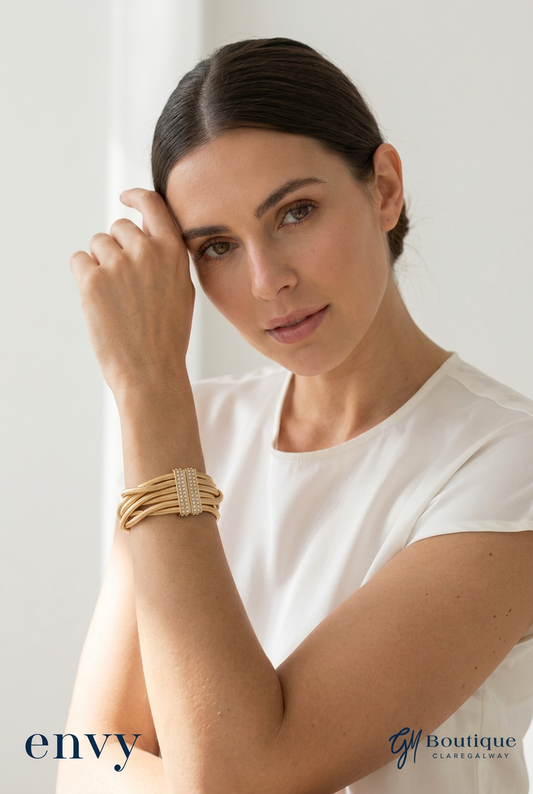 A woman wearing a white top and a multi-strand gold bracelet with a central textured band, posed against a bright white background. The brand logos 'envy' and 'Gh Boutique' are visible in the bottom corners.