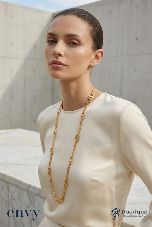 A woman wearing a cream-colored top and a long gold chain necklace with interlocking oval links, posed against a modern concrete wall background. Logos for 'envy' and 'Gm Boutique' are visible in the bottom corners.