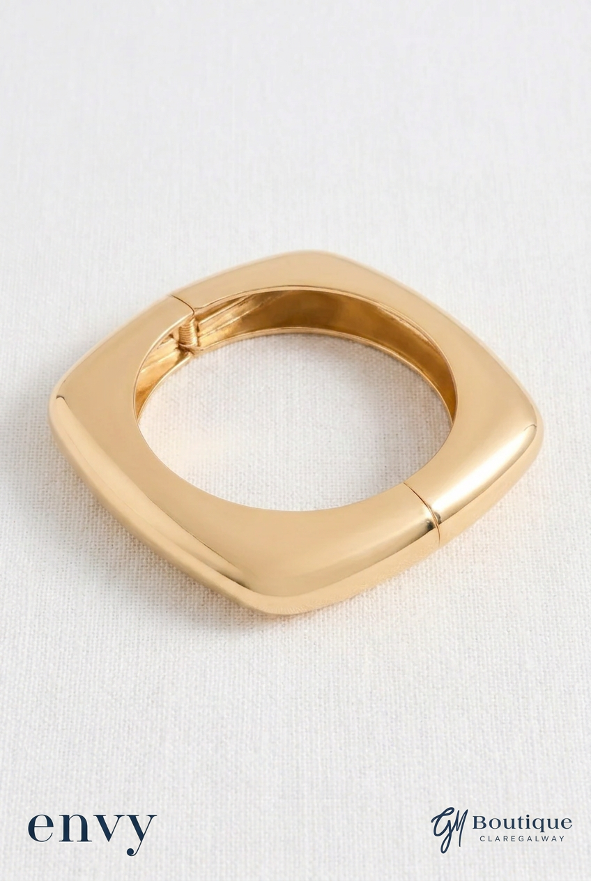 A polished gold square ring with rounded corners, displayed on a textured white fabric background with 'envy' and 'G1 Boutique' brand logos at the bottom.