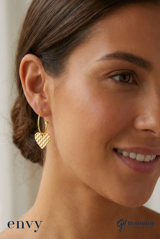 A close-up of a woman wearing a small gold hoop earring with a textured heart dangle. The image features the 'envy' brand logo in the bottom left corner and the 'G1 Boutique' logo in the bottom right.
