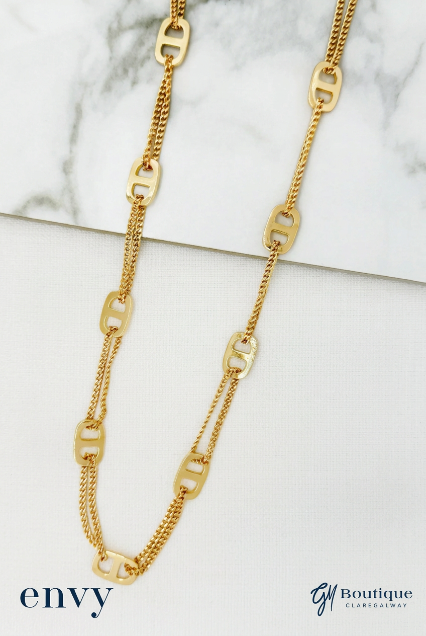 A gold-toned double-strand necklace with elongated oval links and a fine chain, displayed on a white marble and white surface. The brand logos 'envy' and 'Gm Boutique' are visible in the bottom corners.