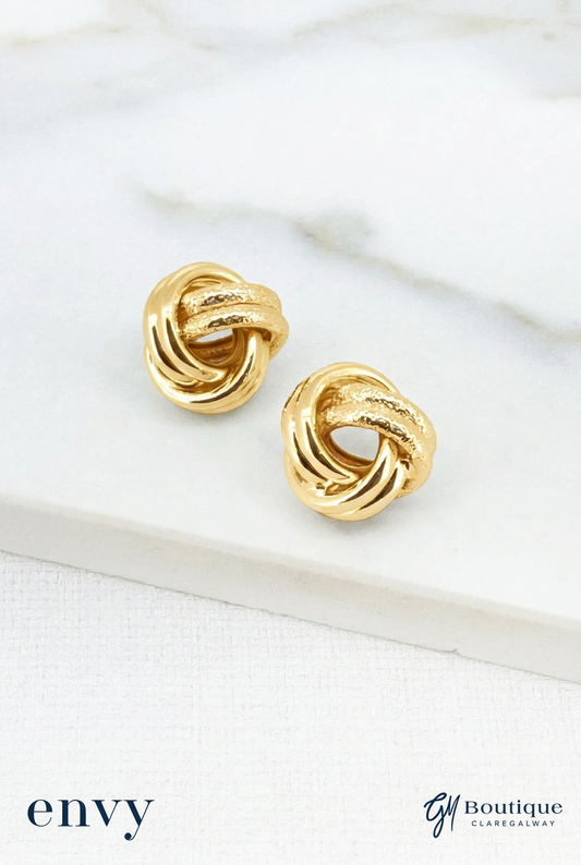 A pair of gold knot stud earrings displayed on a white marble slab over a white textured surface. The brand logos 'envy' and 'Gm Boutique GLAMRAGAWAY' are visible in the bottom corners.