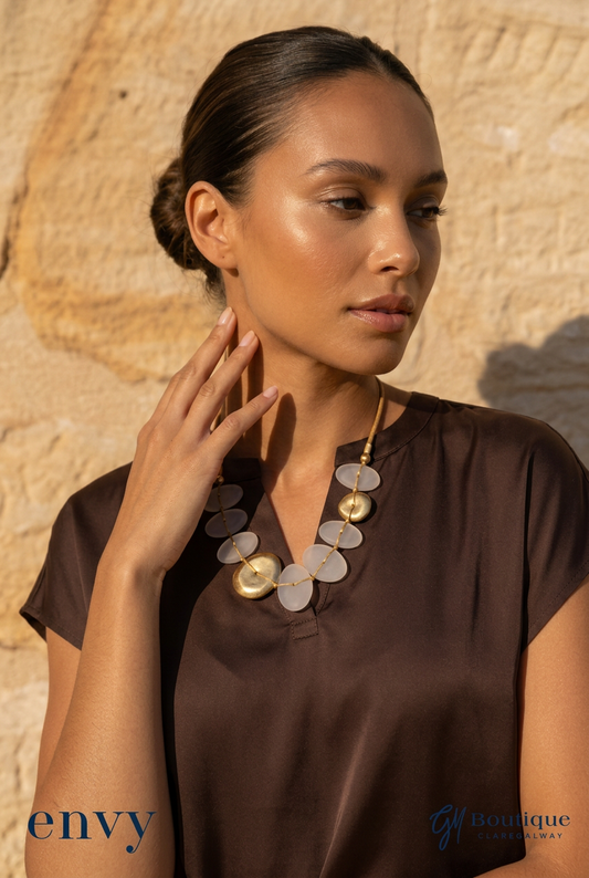 A woman wearing a brown satin blouse and a chunky statement necklace with circular white and gold elements, posing against a textured beige stone wall. The 'envy & GMBoutique' logo is visible in the bottom left corner.