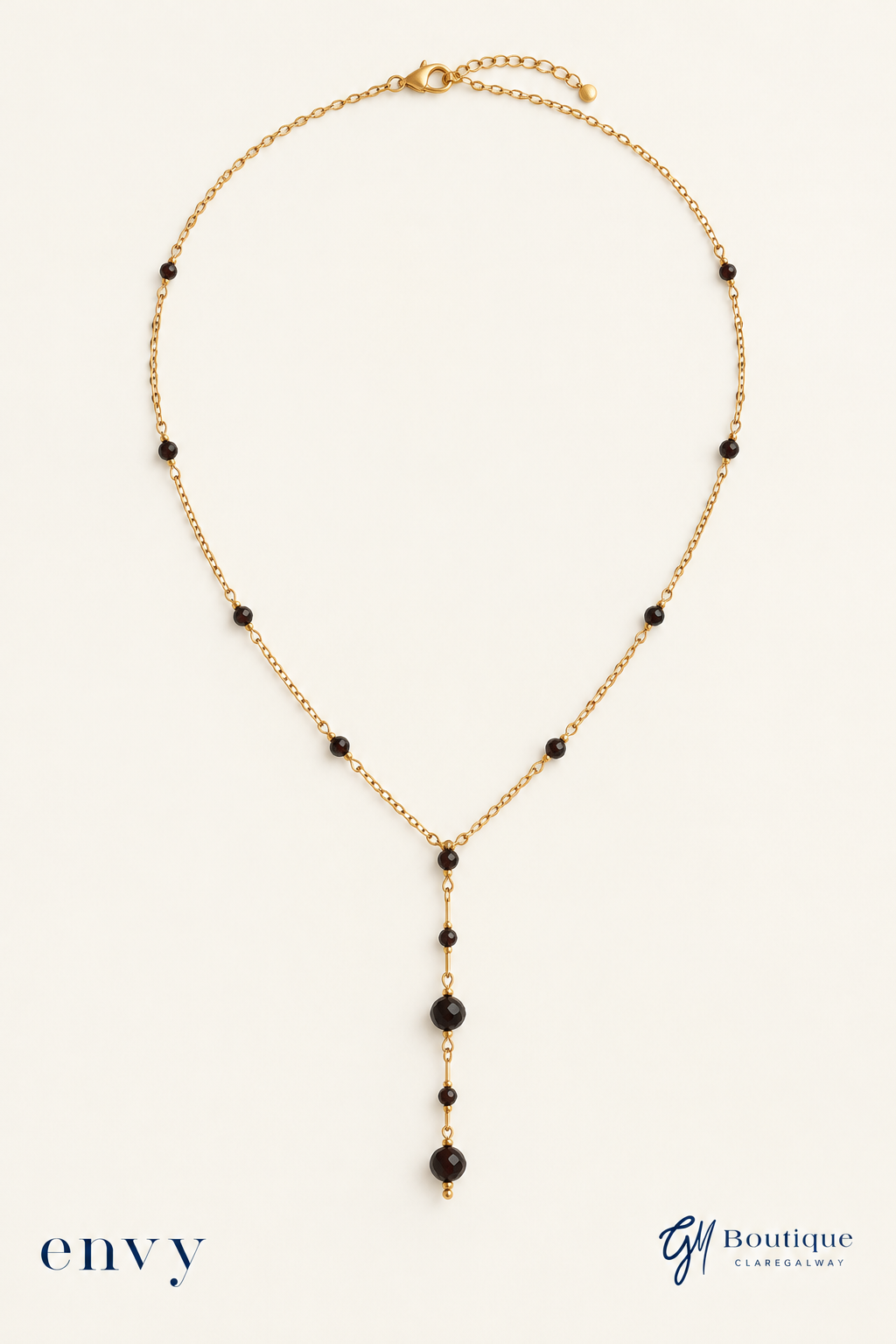 A gold chain necklace with small black beads and a long dangle featuring larger black beads, displayed on a light cream background with 'envy' and 'GM Boutique' branding at the bottom.