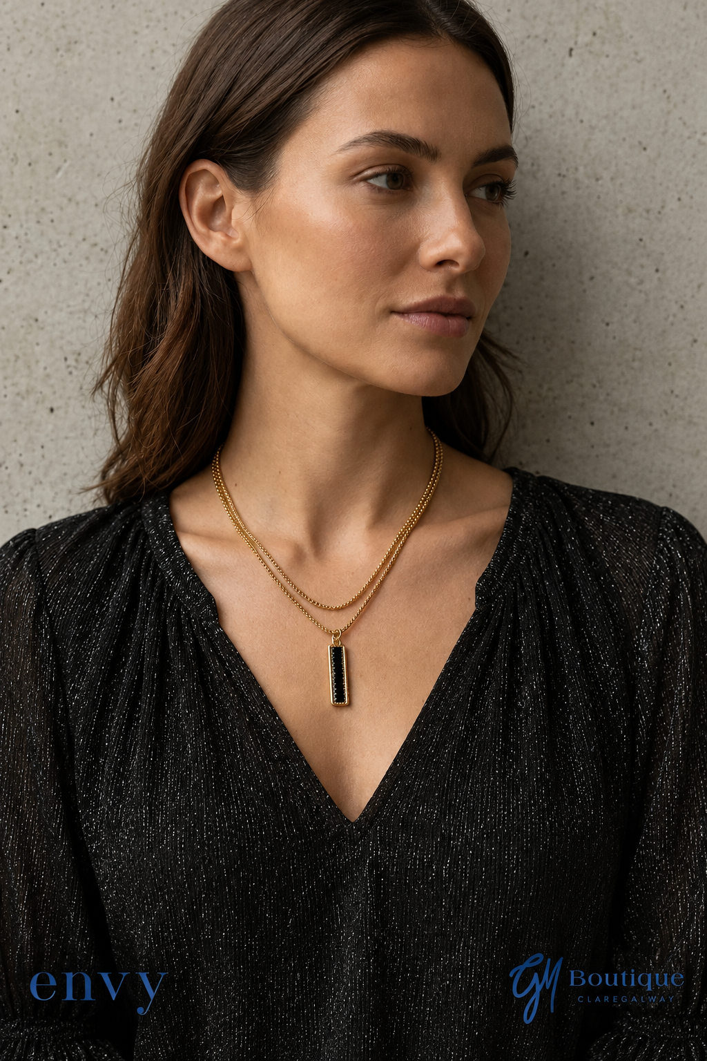A woman wearing a double-layered gold necklace with a vertical black rectangular pendant, styled with a black textured top against a concrete wall background Styled by GM Boutique Galway