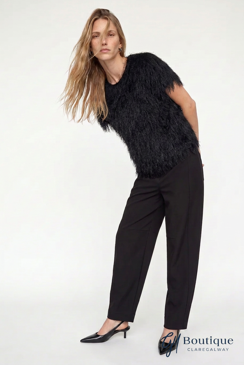 A woman modeling a black feather-trimmed short-sleeve top paired with black wide-leg trousers and black pointed-toe slingback heels, photographed against a plain white studio background.