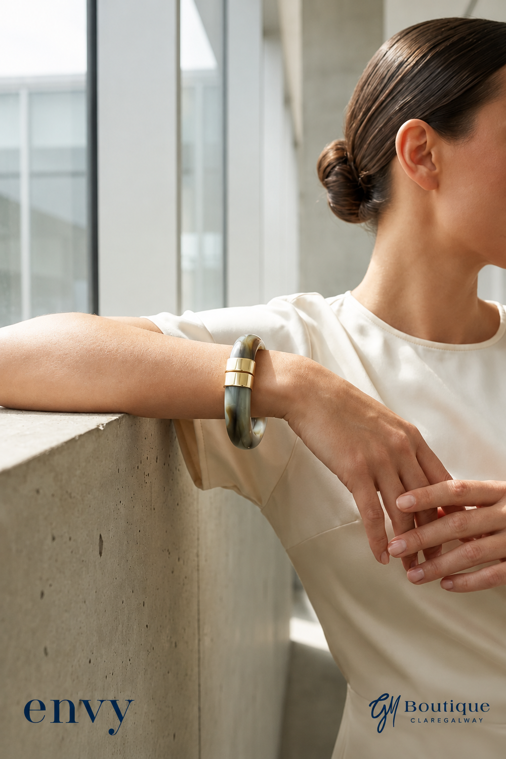 A woman in a cream-colored top leans on a concrete ledge, wearing a thick, dark green jade-like bangle with gold accent bands. The image features 'envy' and 'G1 Boutique' watermarks in the bottom corners.