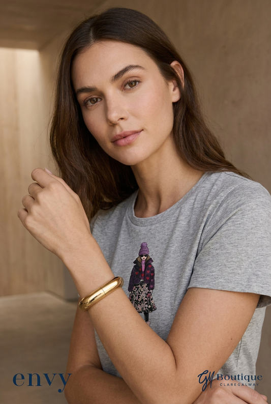 A woman wearing a heather gray t-shirt with a graphic of a woman in a purple coat, accessorized with a gold bangle bracelet. The image has 'envy' and 'GfB Boutique' watermarks at the bottom.