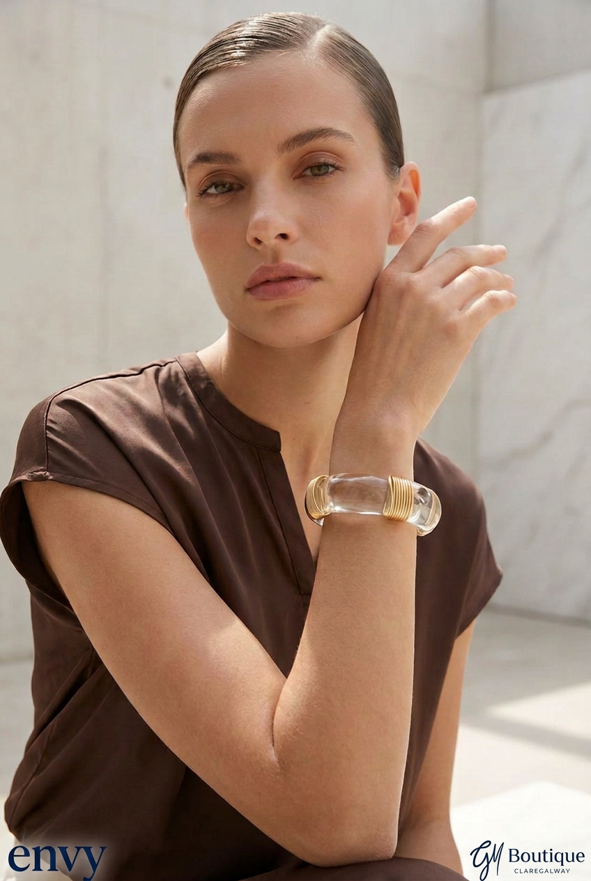 A woman wearing a chunky, clear resin bangle with a gold-toned metal inner lining, posing in a brown top against a light gray marble wall. The 'envy' brand logo is visible in the bottom-left corner.