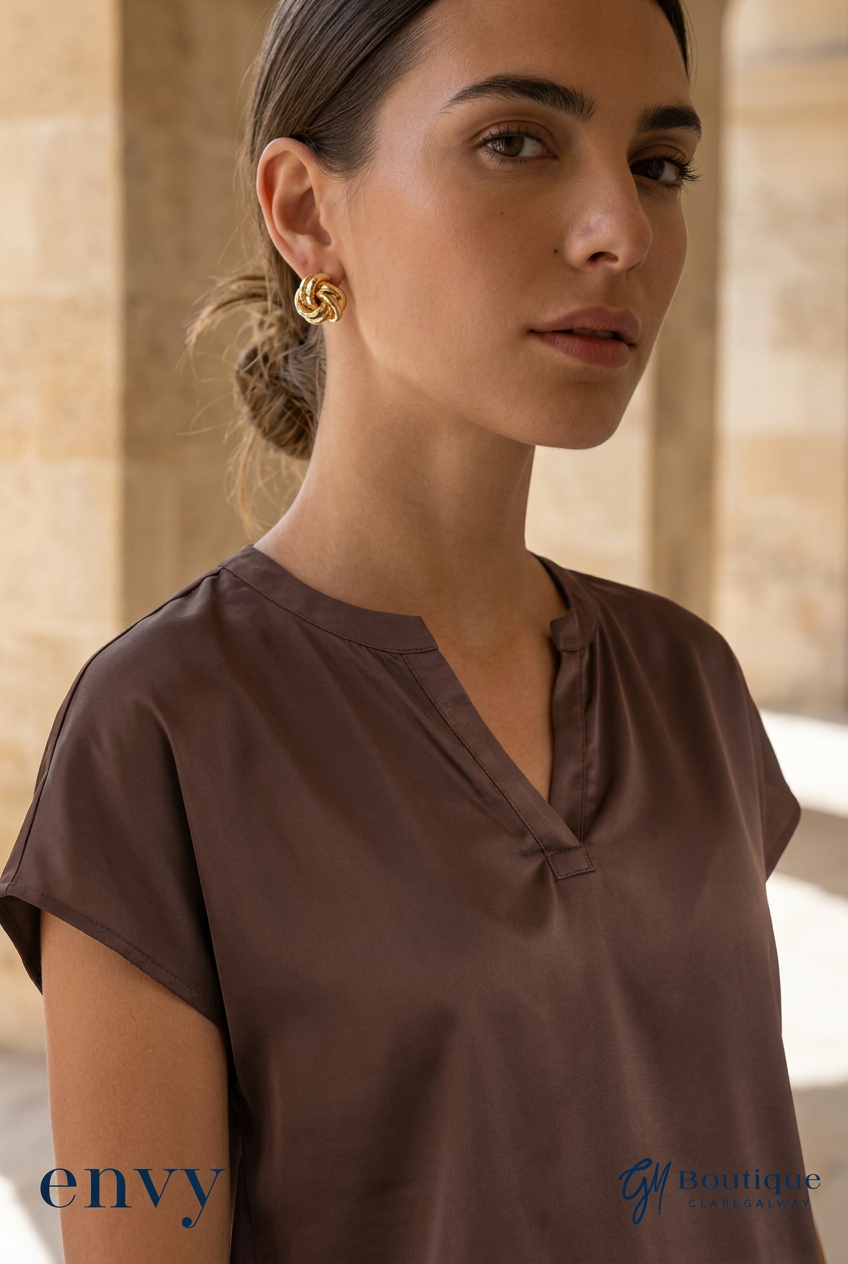 A woman wearing a brown short-sleeve blouse and gold hoop earrings, photographed in a softly lit indoor setting with stone columns. The image includes 'envy' and 'Gm Boutique' text marks at the bottom.