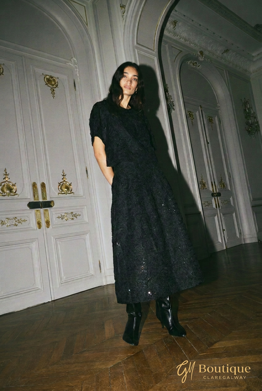 A woman wearing a black textured midi dress with short puff sleeves and black ankle boots, standing in a grand hall with white paneled doors and a dark wood floor.