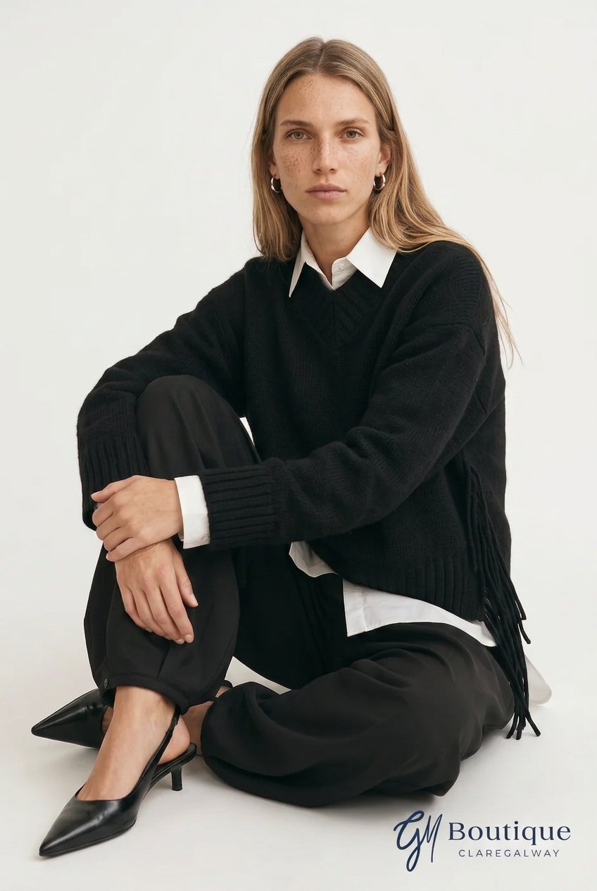 A woman sits cross-legged on a light studio floor wearing a black fringe sweater over a white collared shirt, black trousers, and black pointed heels. A 'GM Boutique Claregalway' logo appears in the bottom right corner.