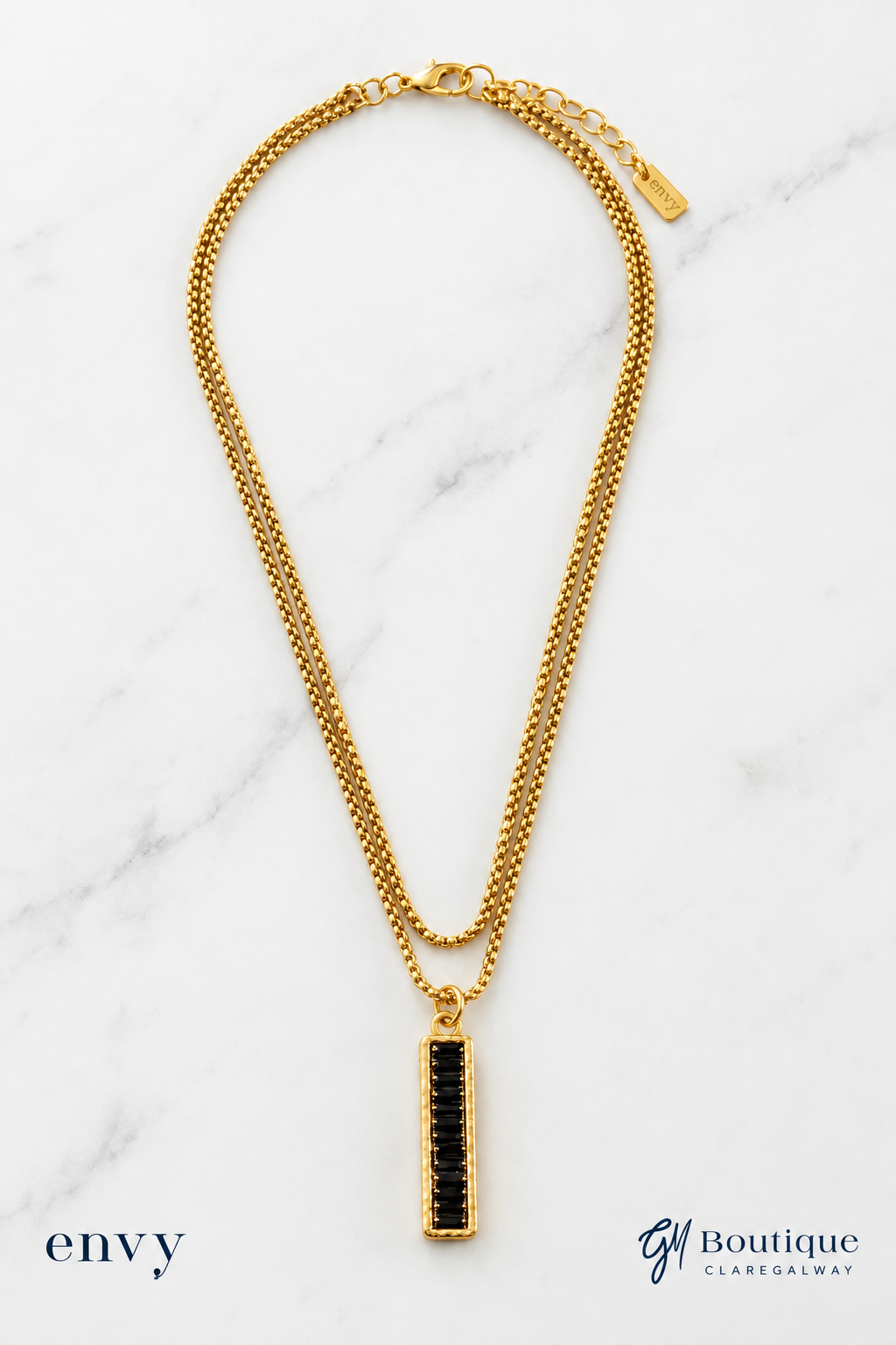 A gold chain necklace with a vertical rectangular pendant set with black stones, displayed on a white marble background with ENVY and GM Boutique Boutique Logos text overlays.