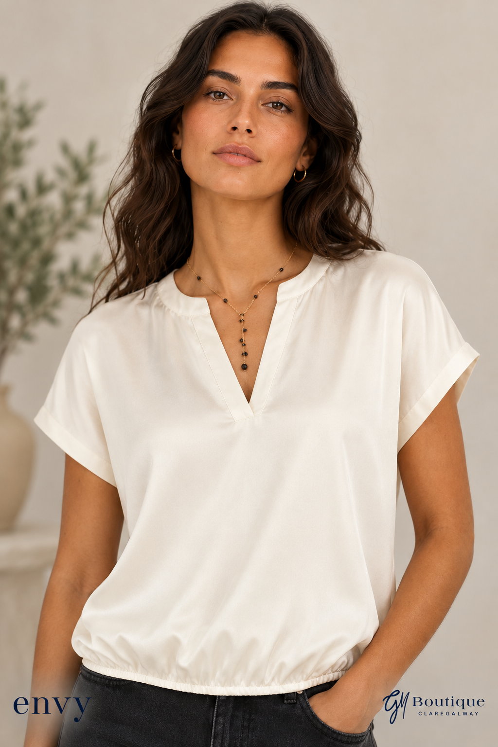 A woman wearing a short-sleeve, off-white satin blouse with a split V-neckline, paired with dark jeans. The photo is taken in a softly lit indoor setting with a neutral background and a plant in the distance.