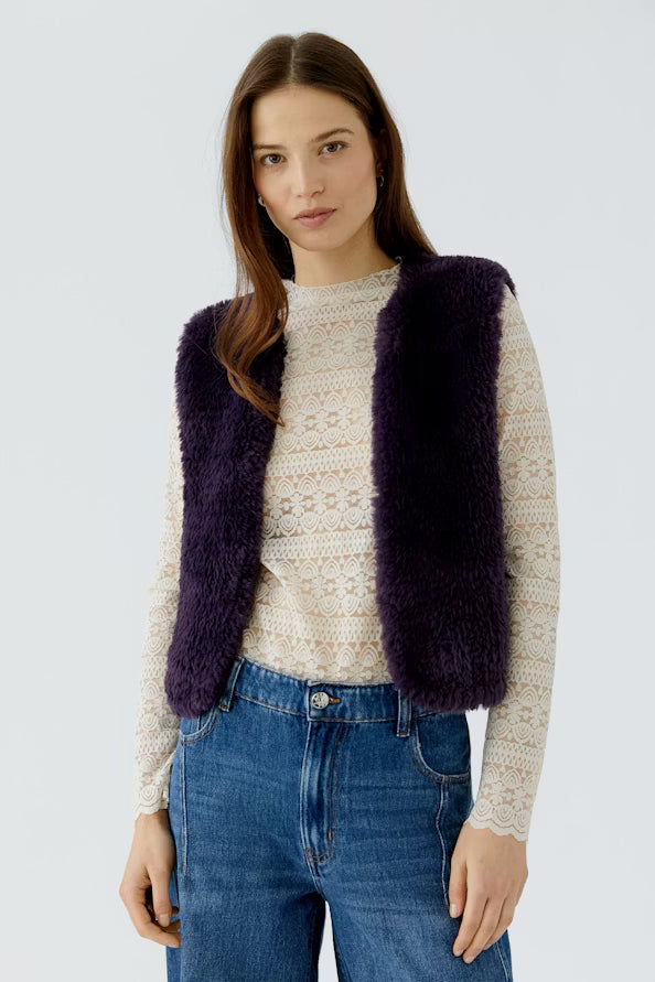 Woman wearing a purple vest over a white sweater with blue jeans on a light gray background