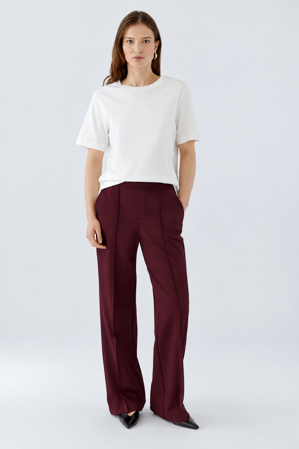 A woman modeling a white short-sleeve crewneck top and dark burgundy wide-leg trousers against a light gray studio background.