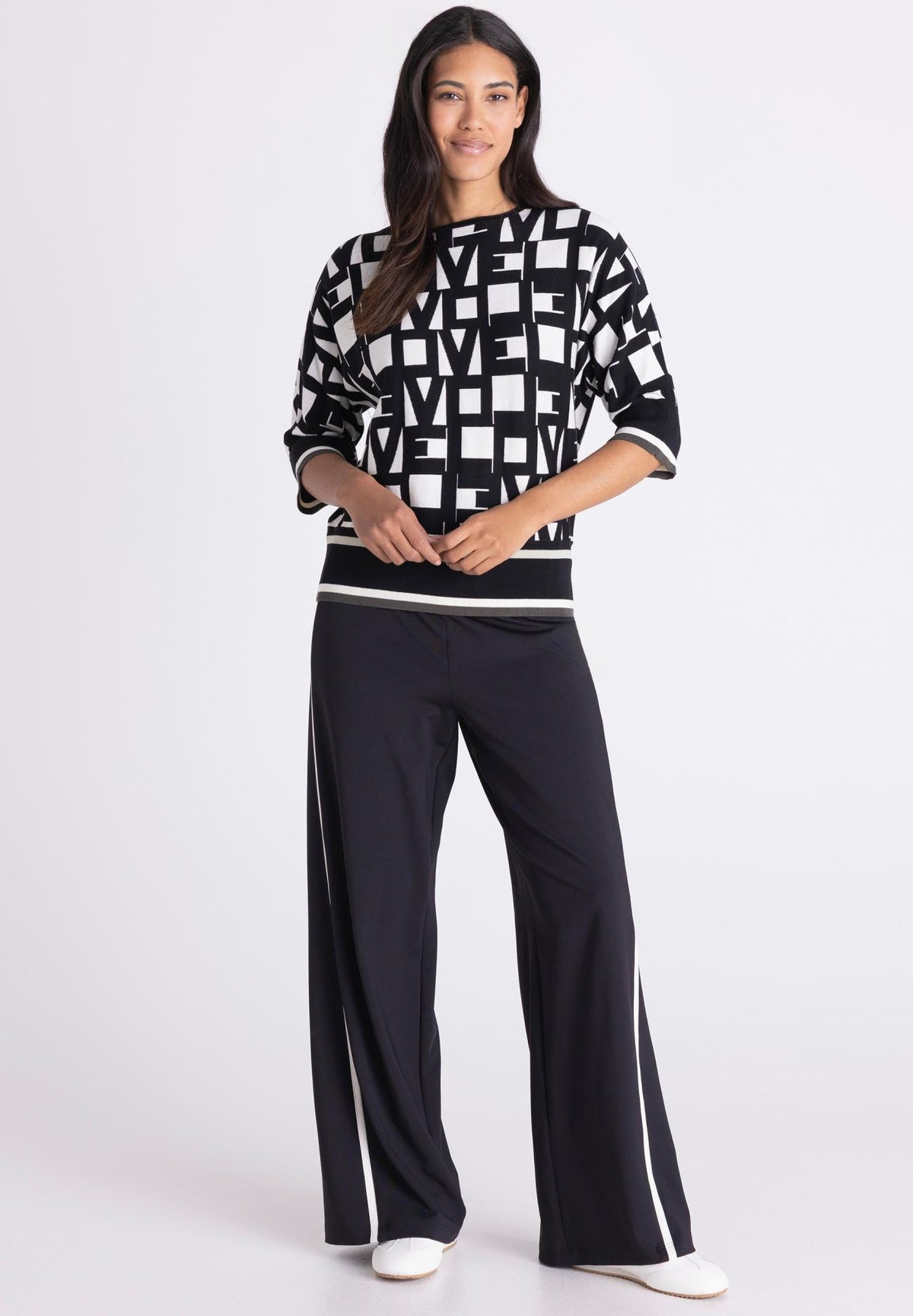 A woman modeling a black and white geometric patterned sweatshirt with three-quarter sleeves and matching black wide-leg pants featuring white side stripes, set against a plain light gray studio background.