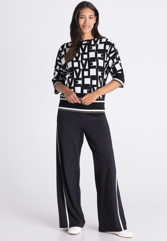 A woman modeling a black and white geometric patterned sweatshirt with three-quarter sleeves and matching black wide-leg pants featuring white side stripes, set against a plain light gray studio background.