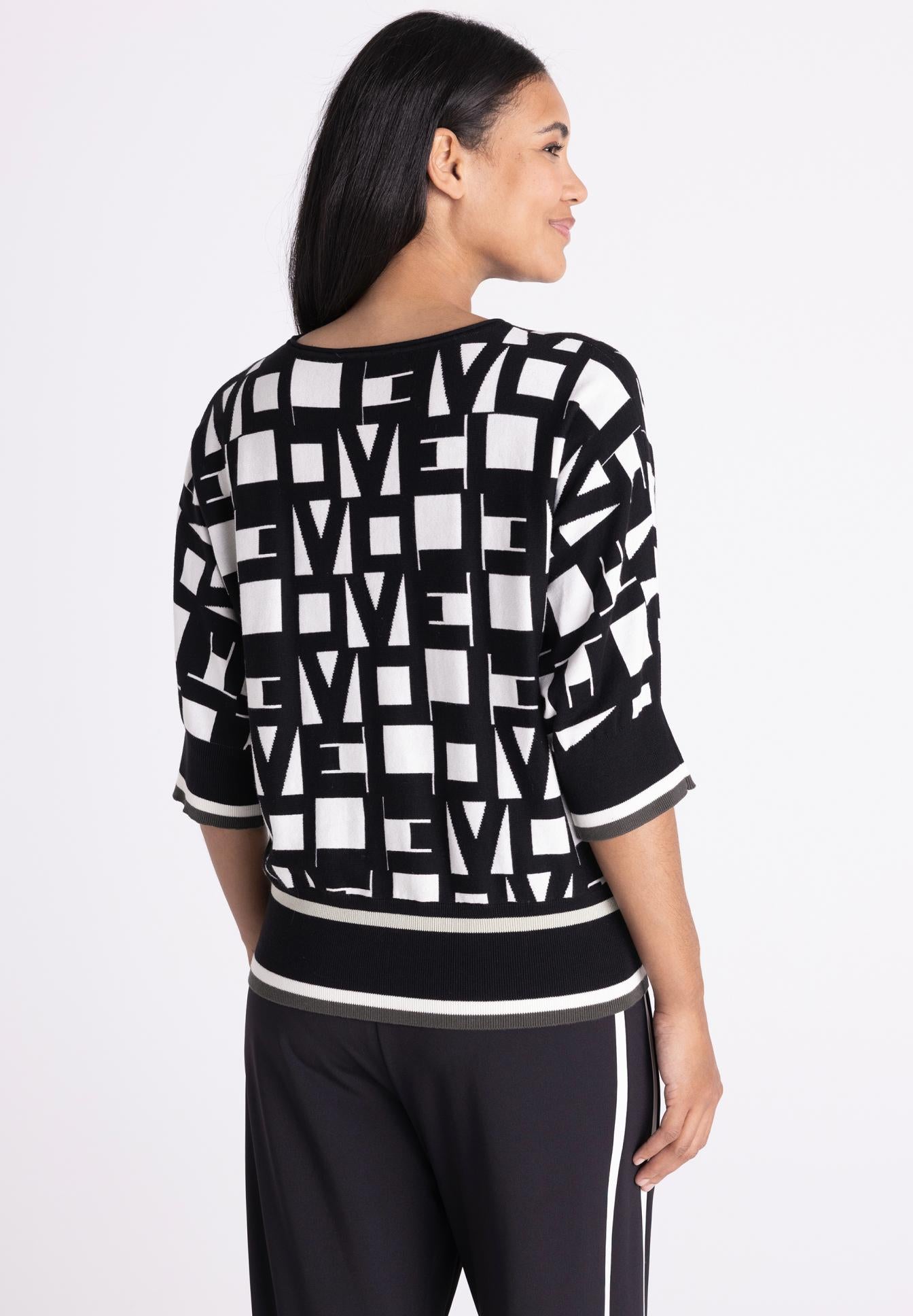 A woman shown from the back wearing a black and white geometric patterned knit sweater with three-quarter sleeves and black knit pants with white side stripes, against a plain light gray studio background.