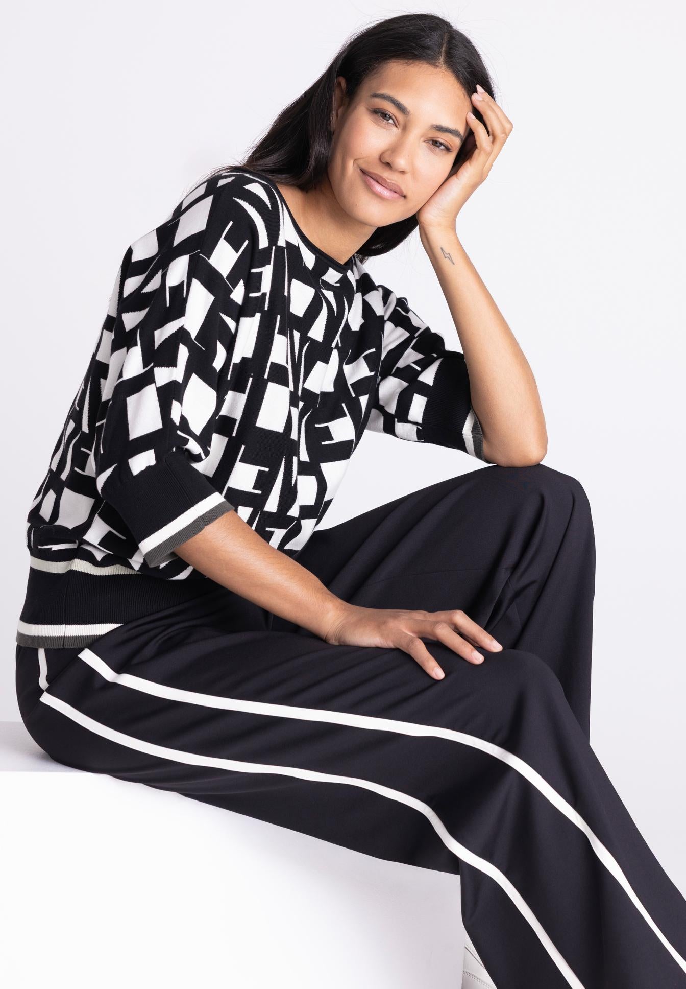 A woman posing in a black and white geometric patterned knit top and matching black wide-leg trousers with white side stripes, set against a clean white studio background.