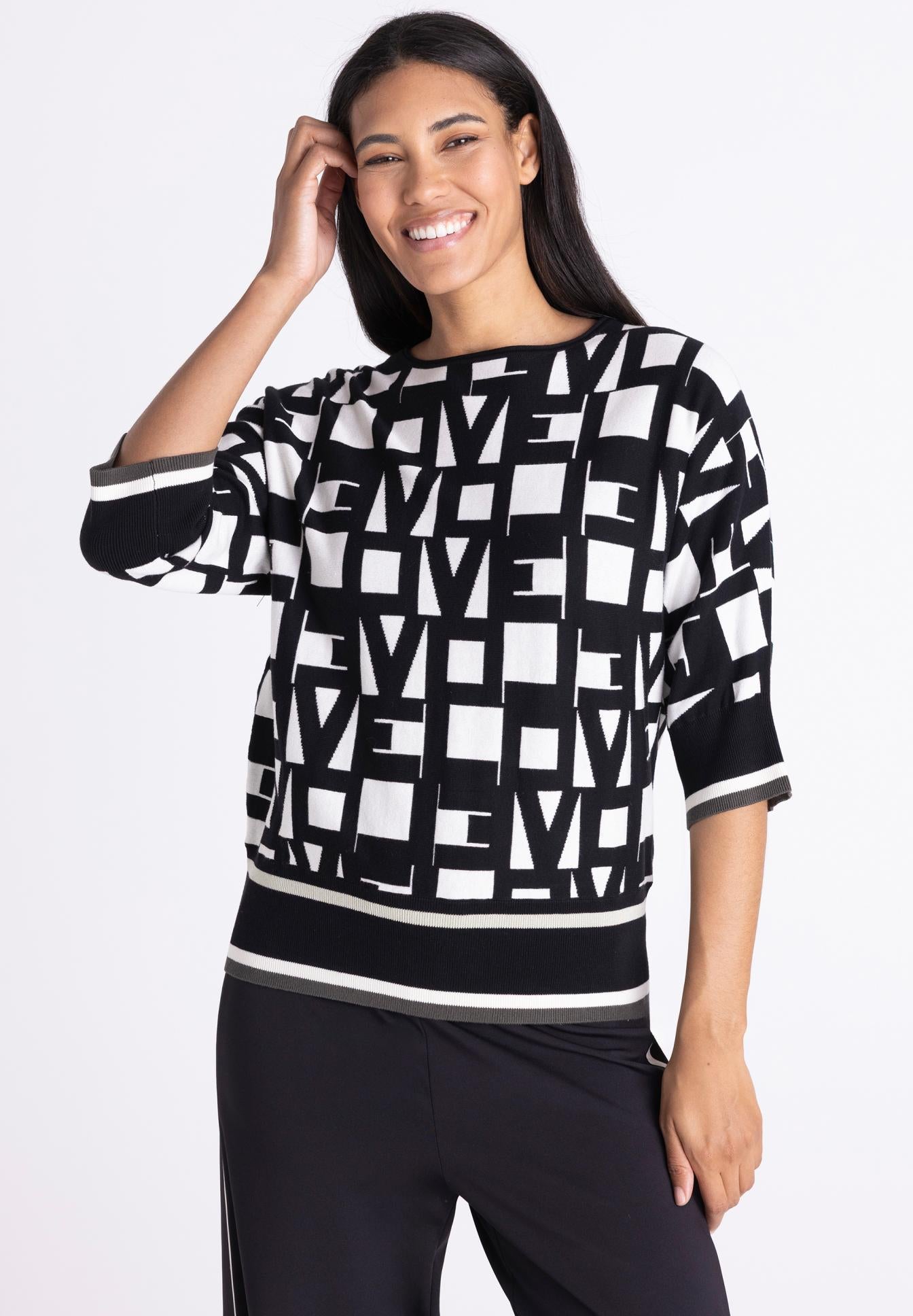 A woman modeling a black and white geometric pattern knit top with three-quarter sleeves and a striped hem, paired with black pants, against a plain white studio background.