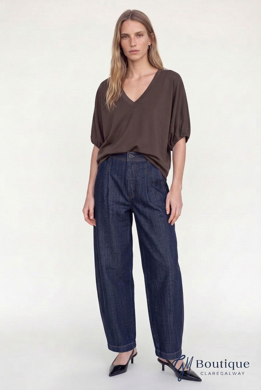 A woman modeling a dark brown short-sleeve V-neck top and dark blue wide-leg jeans against a plain white studio background. A GM Boutique Clarendelway watermark is visible in the bottom right corner.