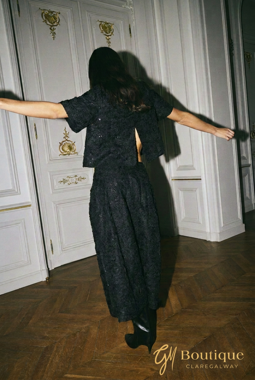 A woman wearing a black textured short-sleeve top and matching maxi skirt, photographed from behind with her arms outstretched in an elegant room with white paneled doors and a wood parquet floor. A 'GM Boutique CLAREGALWAY' watermark is visible in the bottom right corner.