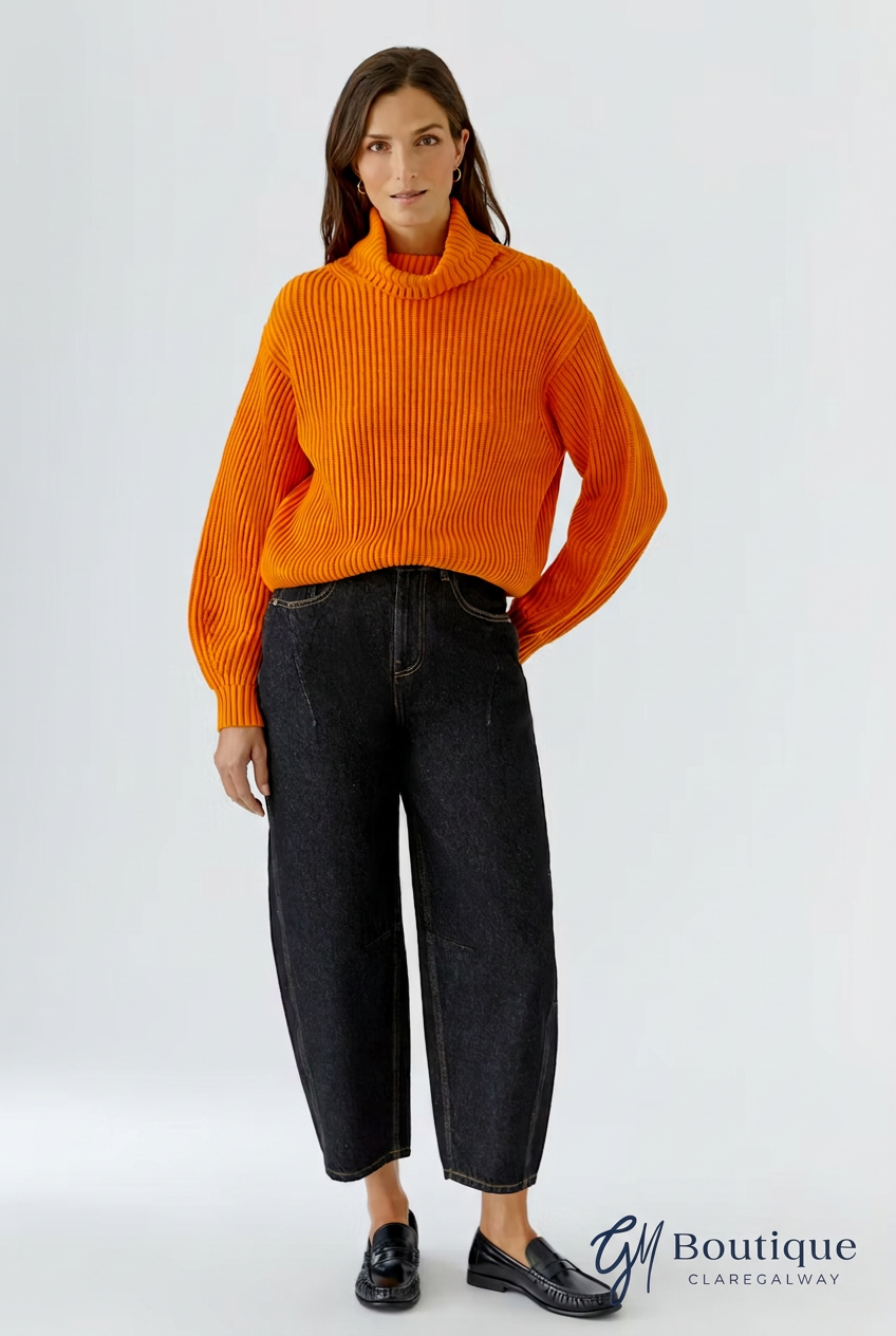 A woman modeling a bright orange ribbed turtleneck sweater paired with dark wide-leg cropped jeans and black loafers, against a light gray studio background. A 'GM Boutique' watermark is visible in the bottom right corner.