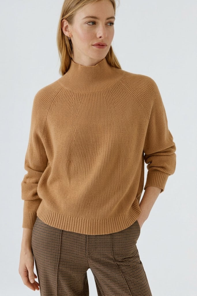 A woman wearing a camel-colored ribbed knit mock neck Oui sweater and brown plaid trousers, photographed against a clean light gray studio background. GM Boutique, Galway