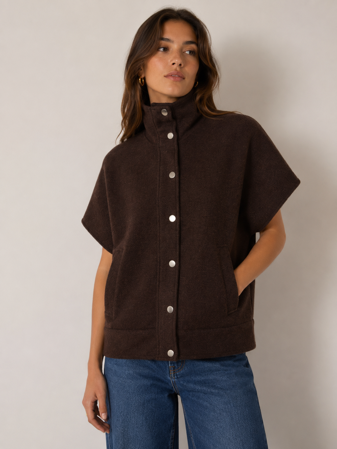 A woman wearing a dark brown short-sleeve button-front jacket with a high mock neck, paired with blue jeans, standing against a plain light gray studio background.