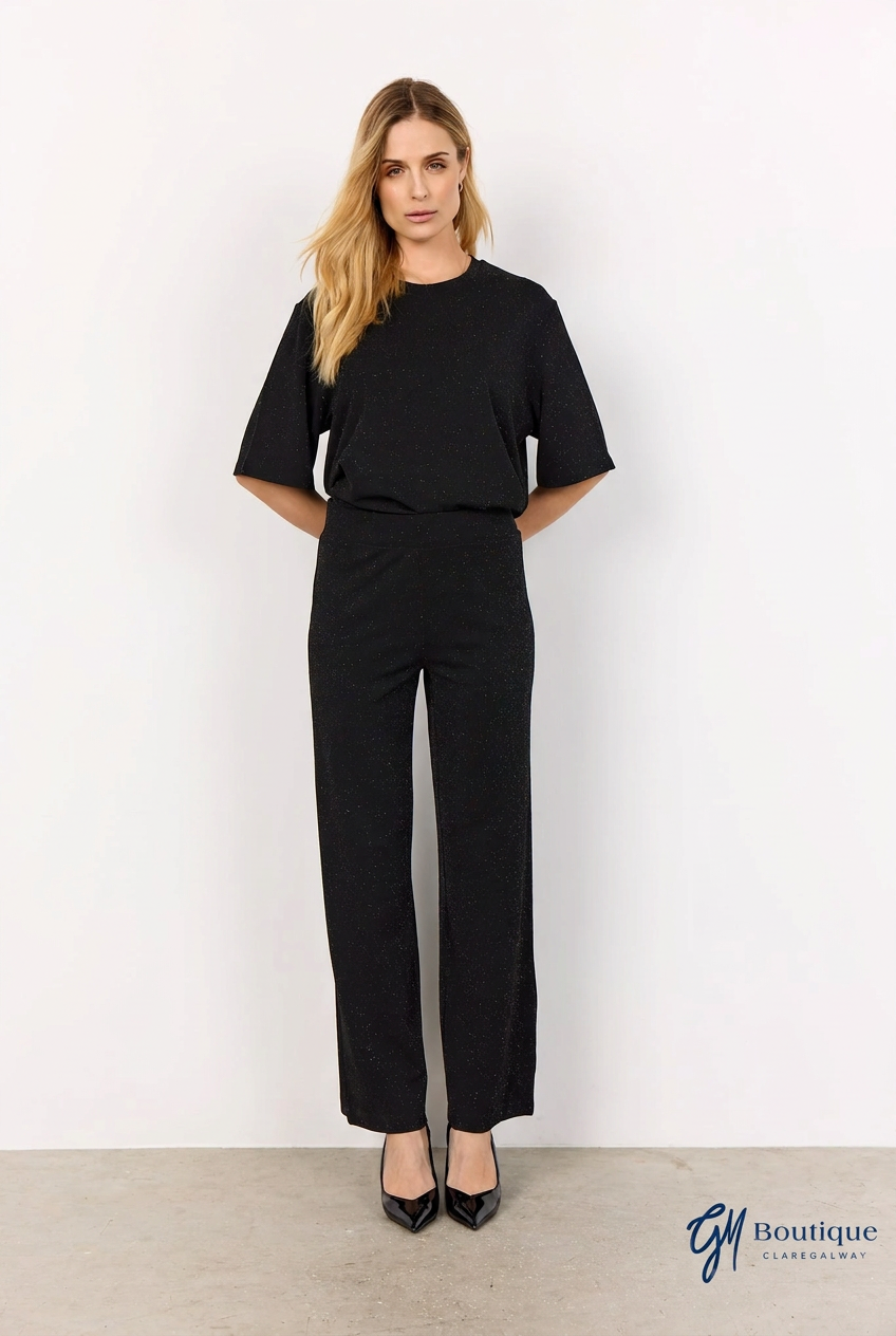 A woman modeling a black short-sleeve top and matching black wide-leg trousers, standing against a plain white wall with a light gray floor. A 'G1 Boutique' logo is visible in the bottom right corner.