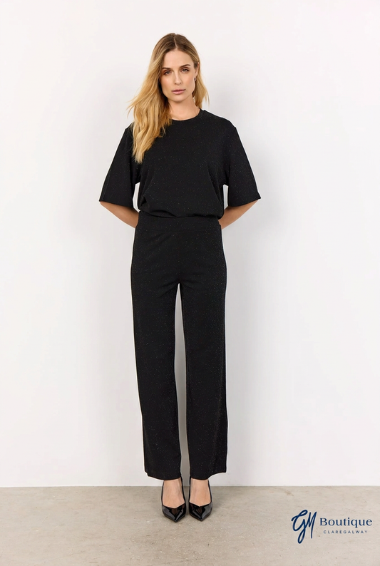 A woman modeling a black short-sleeve top and matching black wide-leg trousers, standing against a plain white wall with a light gray floor. A 'G1 Boutique' logo is visible in the bottom right corner.