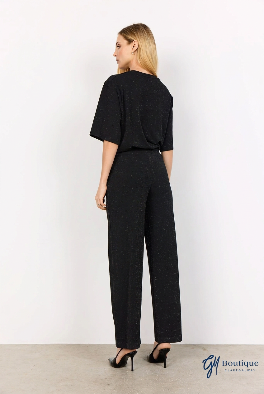 A woman shown from the back wearing a black short-sleeve jumpsuit with wide legs and black high heels, standing against a plain white wall.