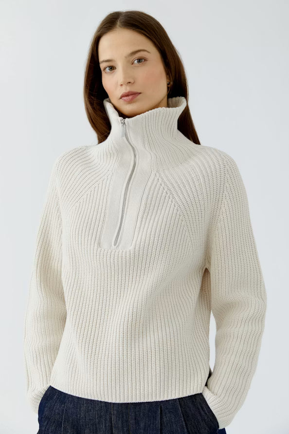 A woman wearing a cream-colored ribbed knit mock-neck sweater with a half-zip closure, paired with dark blue pleated bottoms, against a light gray studio background.