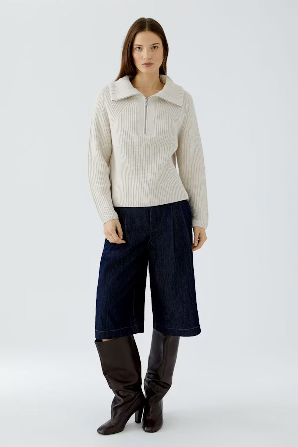 A model stands against a light gray studio background wearing a cream ribbed half-zip sweater, dark blue cropped denim pants, and dark brown knee-high boots.