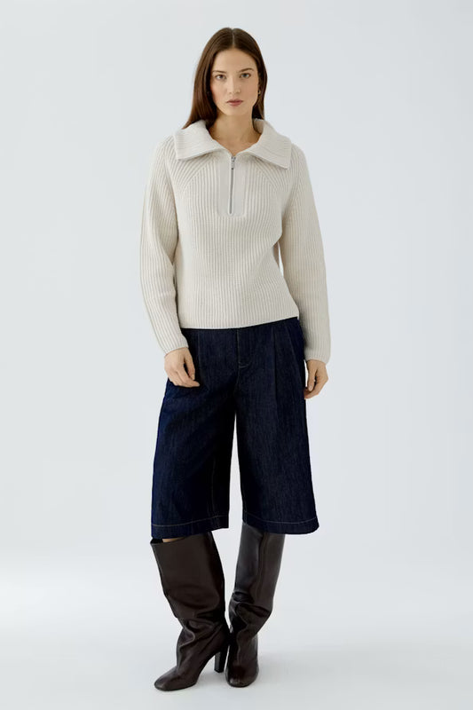 A model stands against a light gray studio background wearing a cream ribbed half-zip sweater, dark blue cropped denim pants, and dark brown knee-high boots.