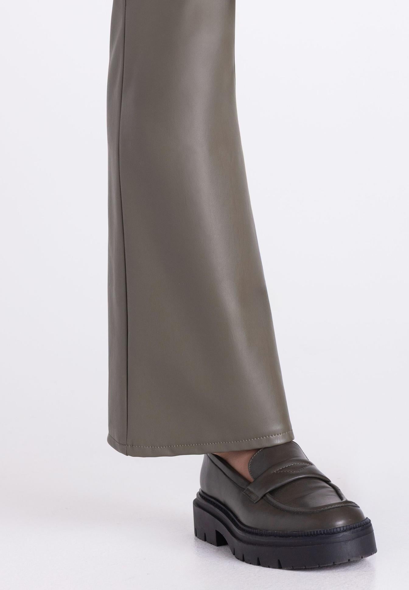 A close-up of a person's leg wearing wide-leg taupe faux-leather trousers and a dark olive green platform penny loafer on a white studio background.