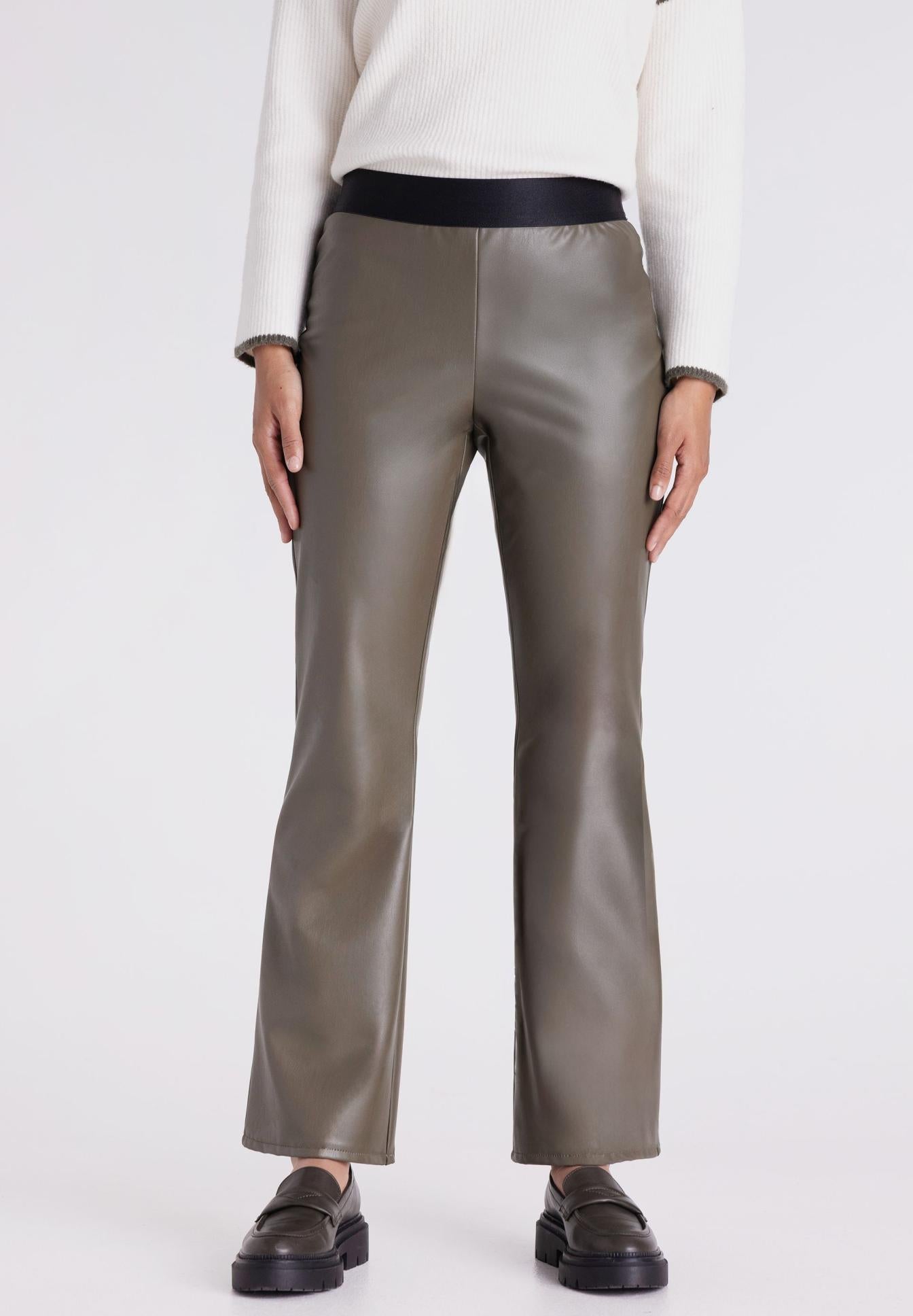 A model is shown from the waist down wearing a pair of olive green faux-leather trousers with a contrasting black waistband, paired with a white sweater and black loafers, against a plain light gray background.