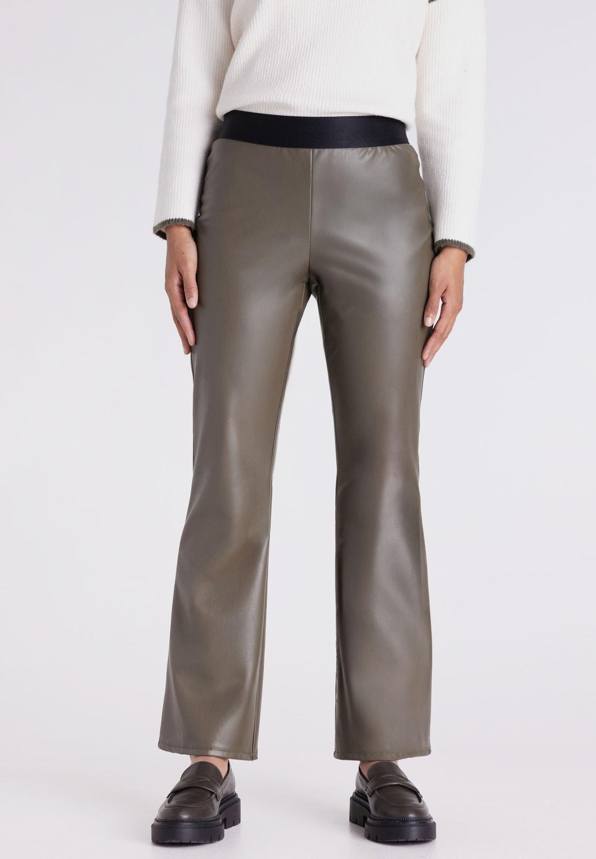 A model is shown from the waist down wearing a pair of olive green faux-leather trousers with a contrasting black waistband, paired with a white sweater and black loafers, against a plain light gray background.