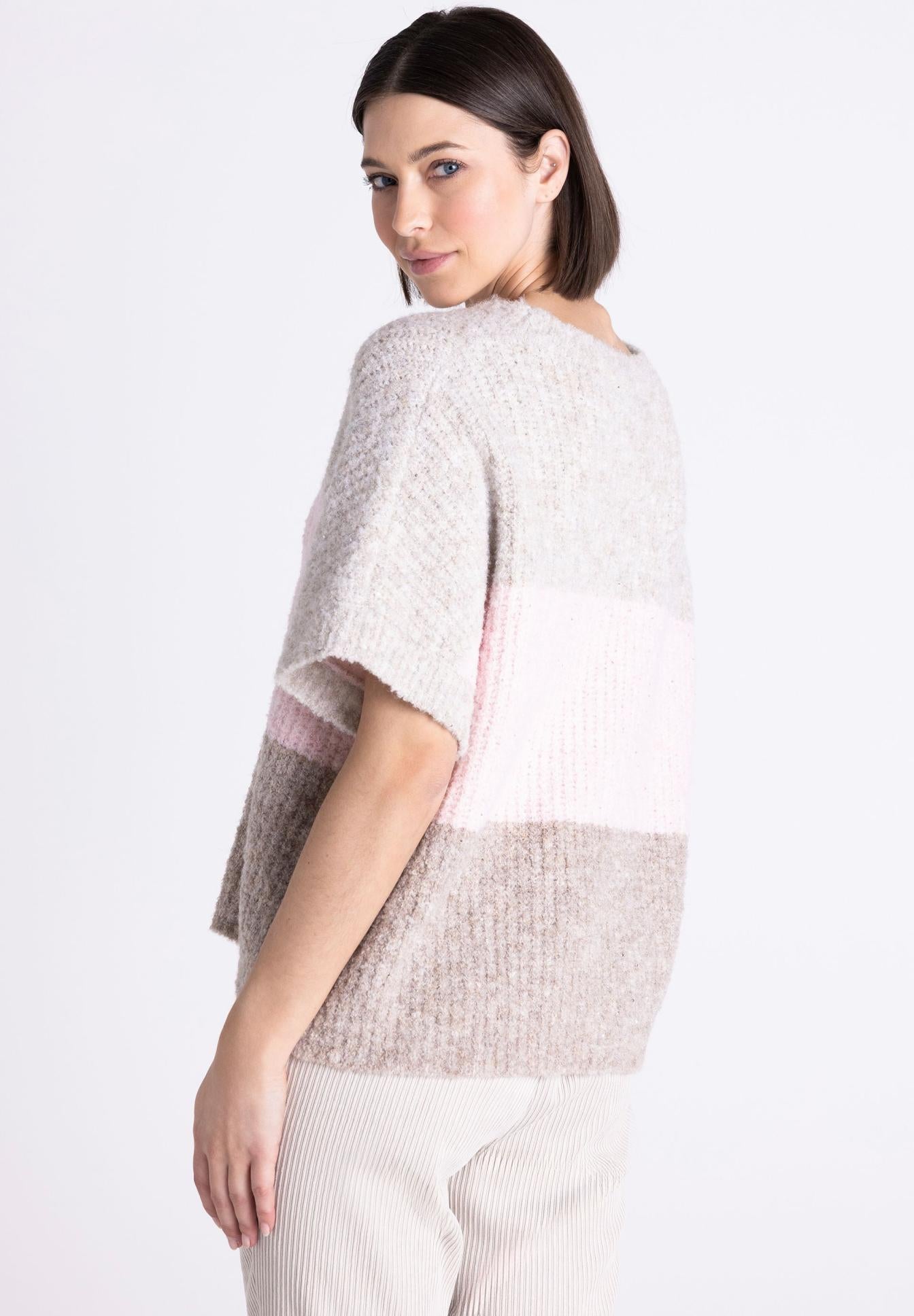 A woman wearing a chunky knit color-block sweater in light gray, pale pink, and taupe, paired with cream ribbed trousers, photographed against a clean white studio background.
