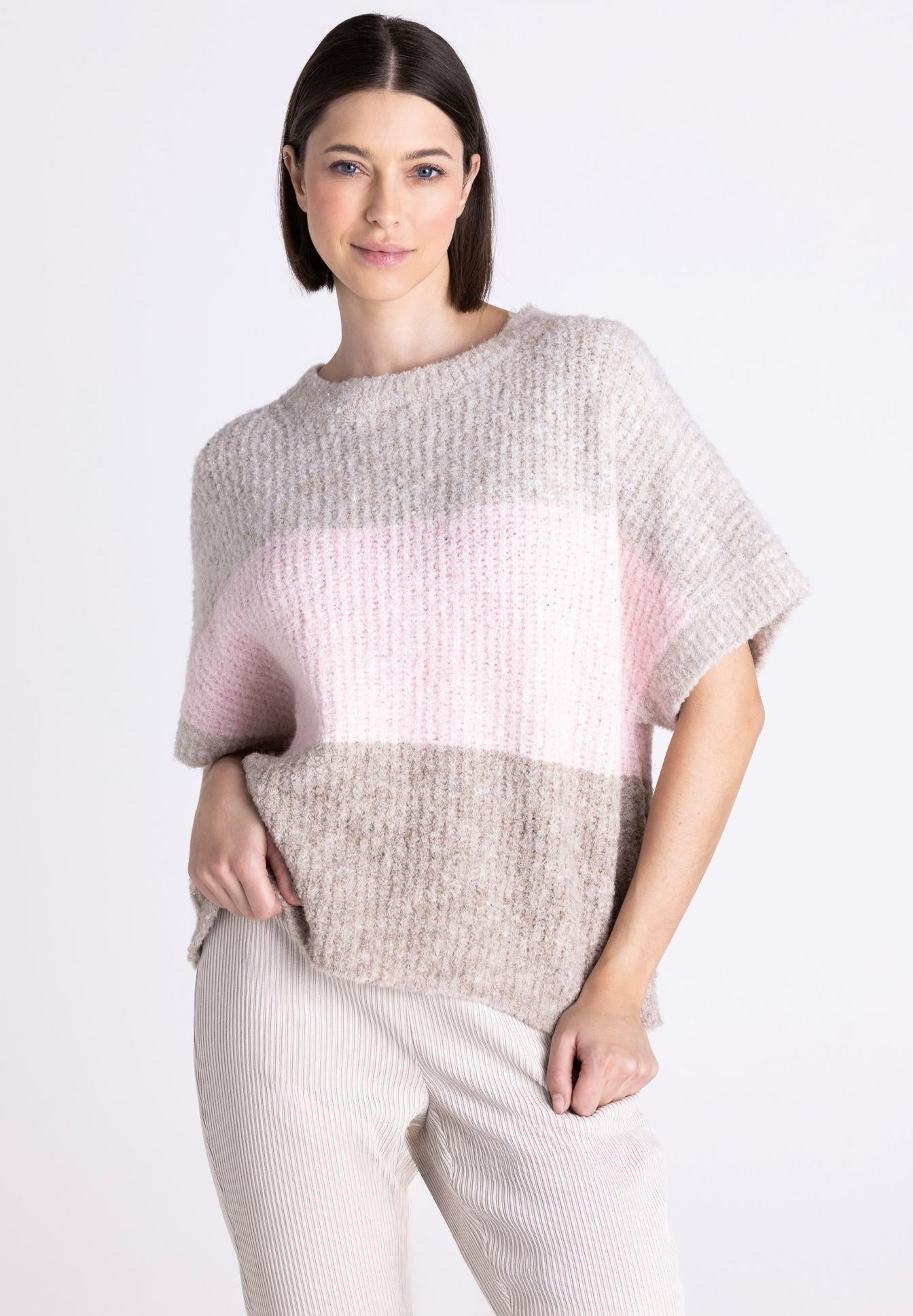 A woman modeling a chunky knit short-sleeve sweater with horizontal color-blocked stripes in light gray, soft pink, and taupe-beige, paired with light beige ribbed pants against a plain white studio background.