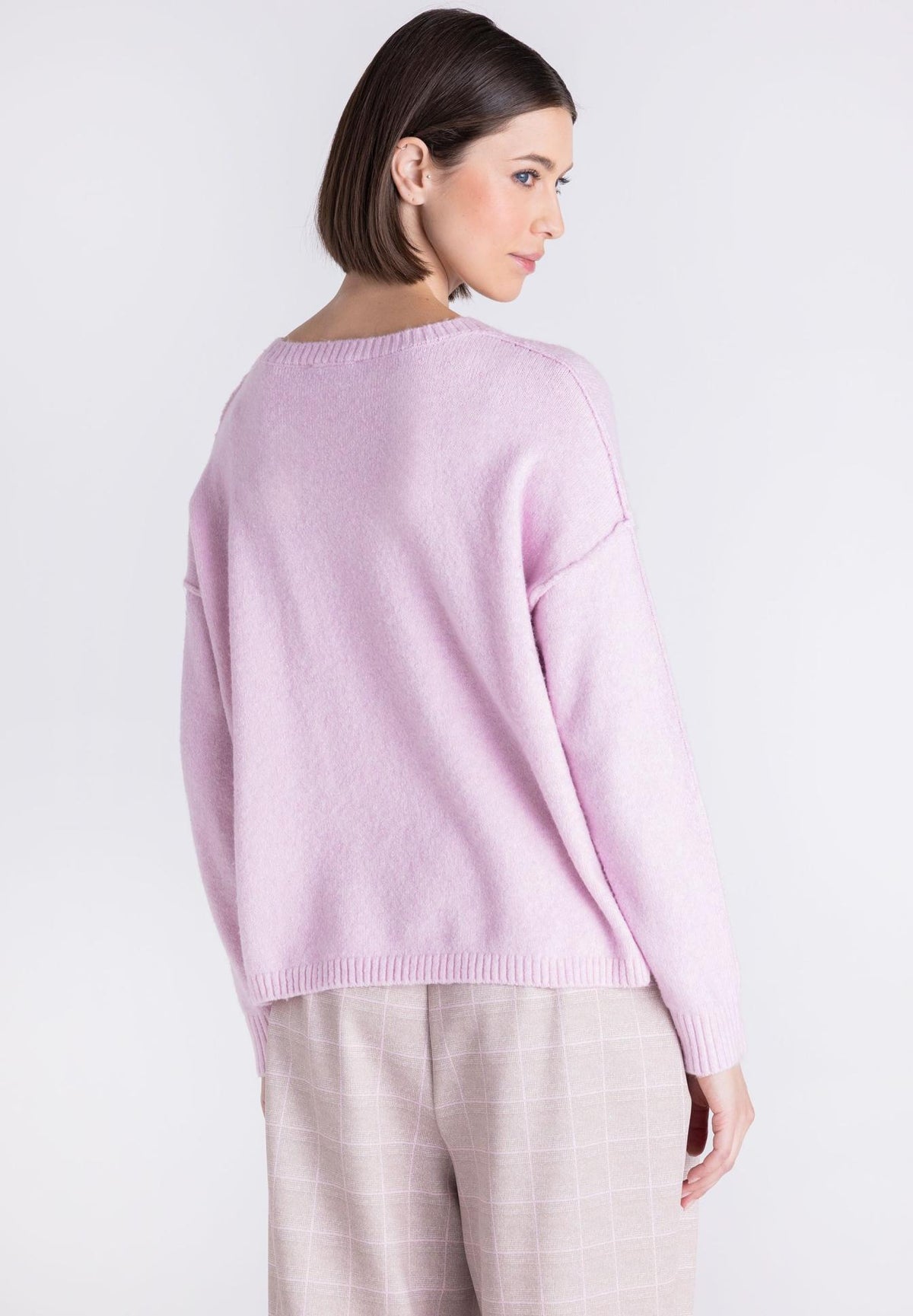 A woman shown from the back wearing a light pink knit sweater and light beige plaid trousers against a plain light gray studio background.